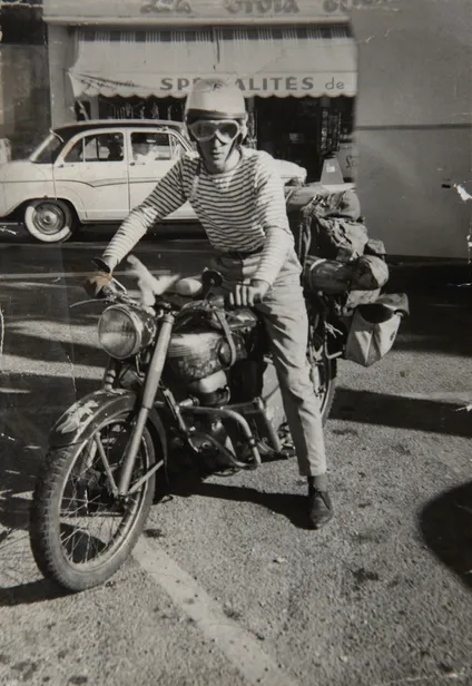 Photo en noir et blanc, homme casque sur la tête, t-shirt marin rayé, pantalon sur sa moto, au fond, une boutique et une vieille voiture.